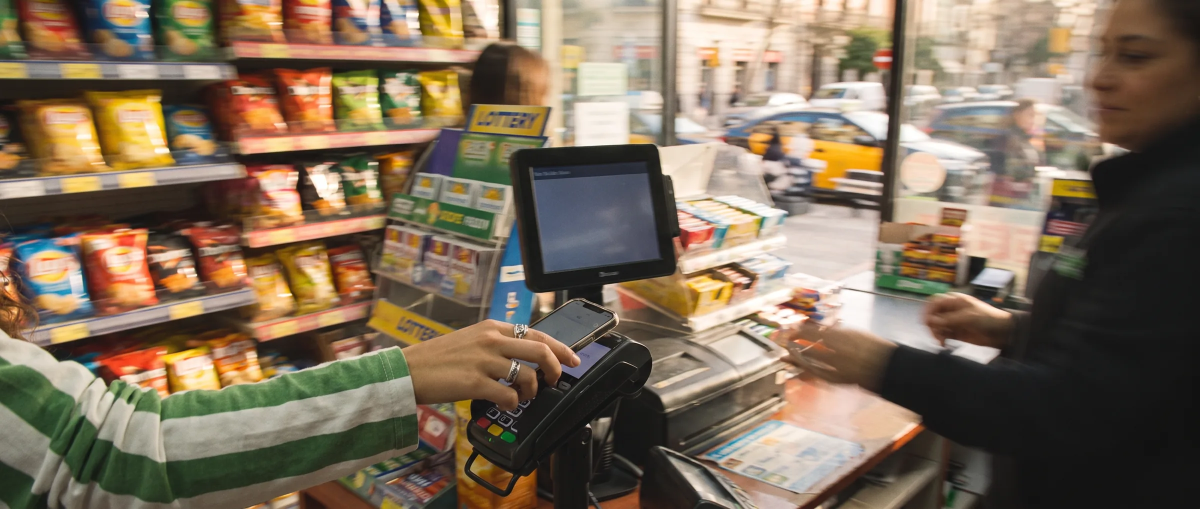 A hand tapping a phone to pay at a bodega counter while the street streaks past.